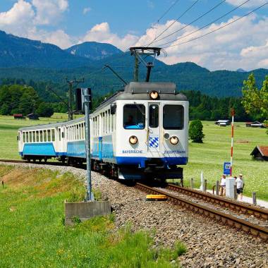 Die Zugspitzbahn fährt auf den Gleisen, im Hintergrund erstrecken sich die Berge.