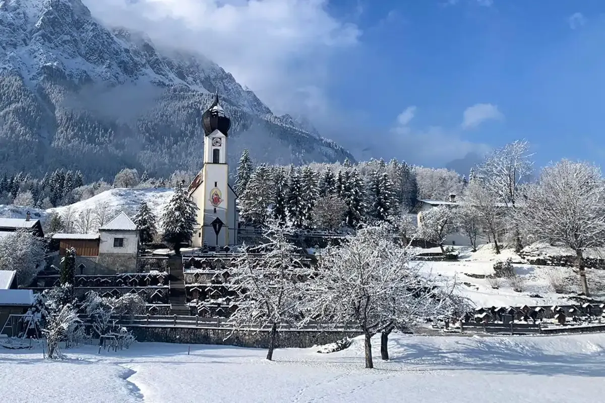 Winterliche Landschaft mit einer Kirche in der Mitte, umgeben von Bergen und Tannen im Hintergrund.