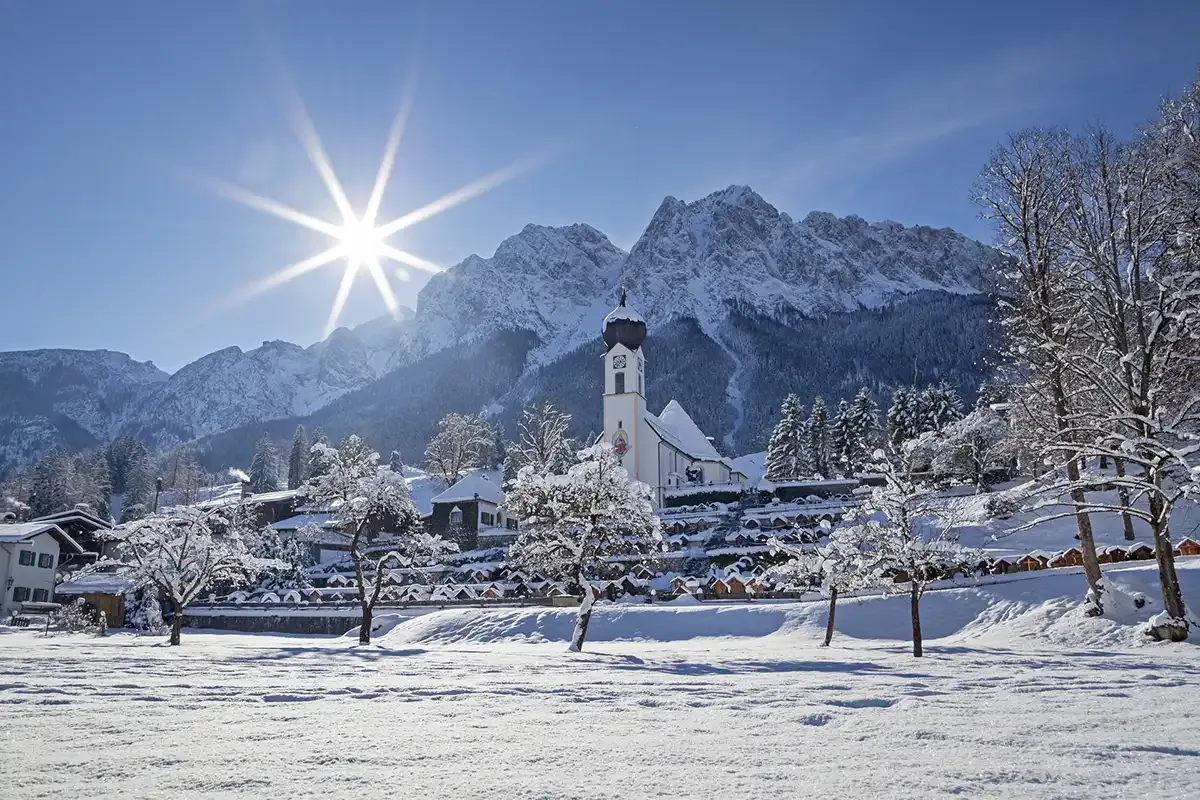 Winterlandschaft in den Alpen mit schneebedeckten Bäumen und Gebäuden. Im Hintergrund eine Kirche und Berge.