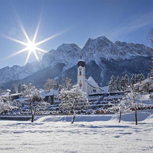 Schneebedeckte Gebirgslandschaft an einem sonnigen Tag mit einer Kirche und Bergen im Hintergrund.