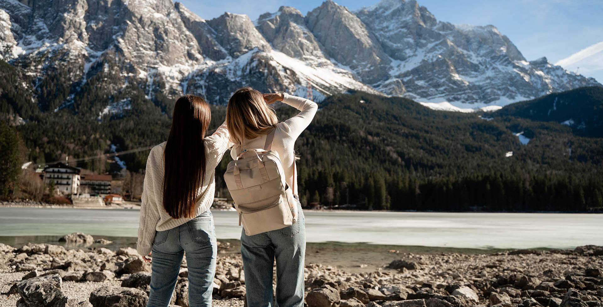 Zwei Frauen stehen am Ufer eines Sees und blicken auf die majestätischen Berge Grainaus in der Ferne.