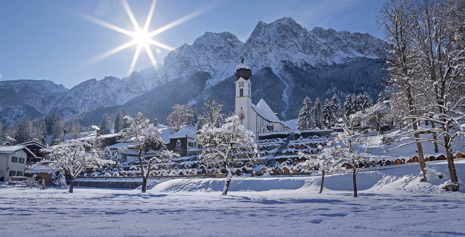 Winterliche Landschaft der bayerischen Alpen mit einer schneebedeckten Ortschaft und Kirche im Hintergrund.