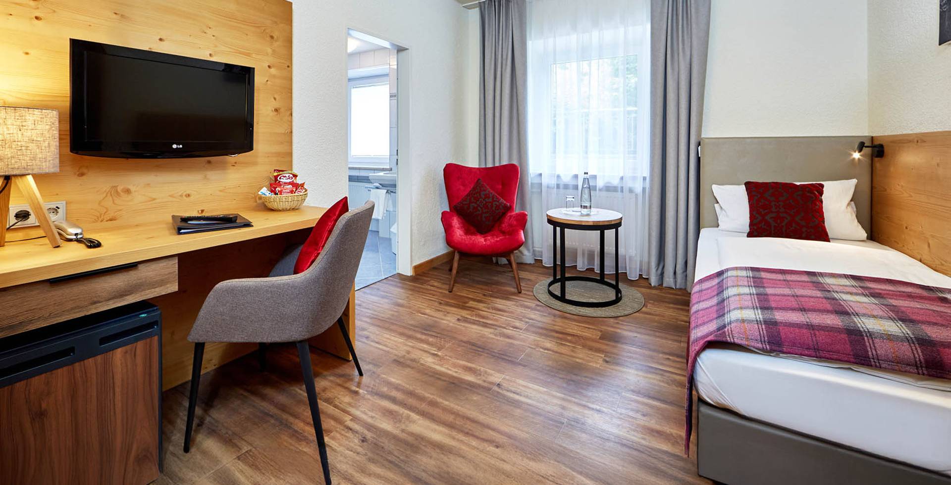 Geräumiges und helles Einzelzimmer mit Einzelbett, TV, Schreibtisch und Kühlschrank mit Blick auf die Berglanschaft.
