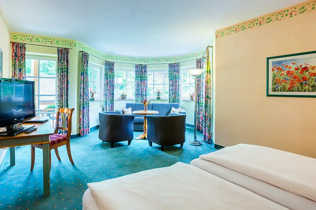 Großes Hotelzimmer im Berghotel Hammersbach mit gemütlicher Sitzecke an einer Fensterfront und Schreibtisch mit Fernseher.
